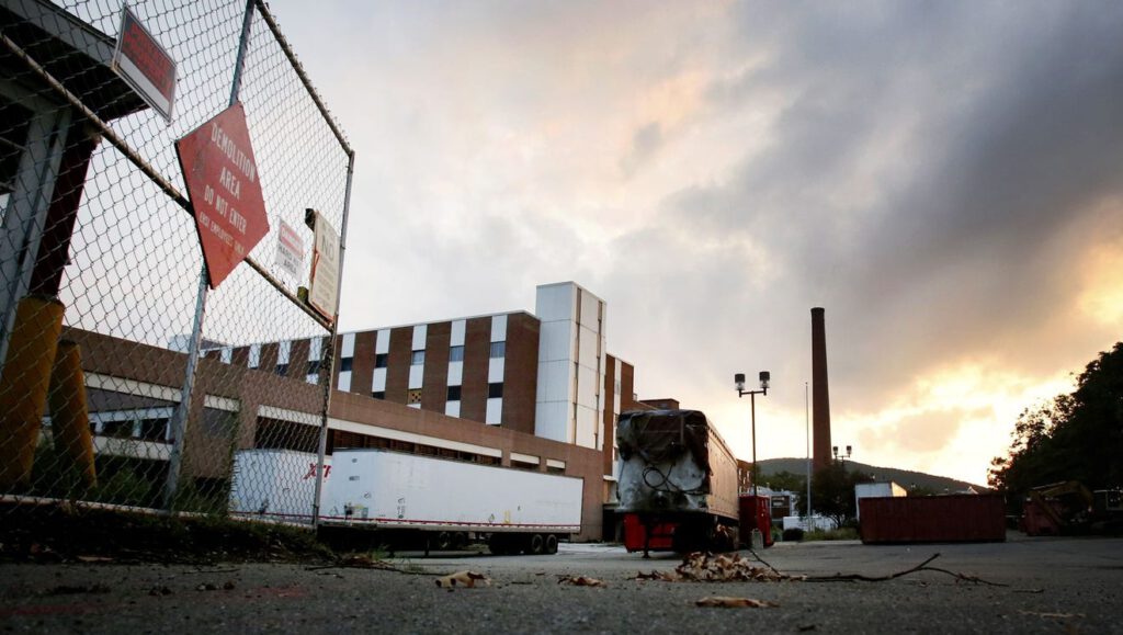 Industrial building with large chimney and trailers, viewed from a fenced area with a warning sign, under a cloudy sky during sunset.