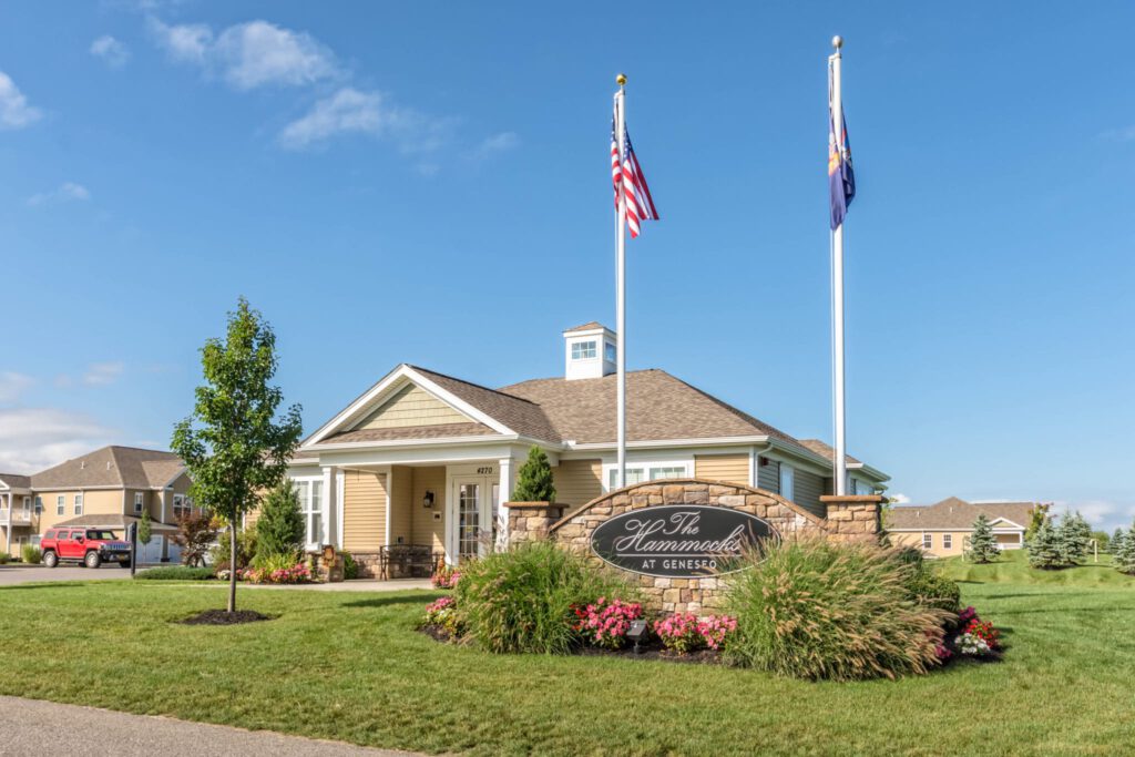 Single-story community center with flags, manicured lawn, and sign reading "The Hammocks at Geneseo" in front.