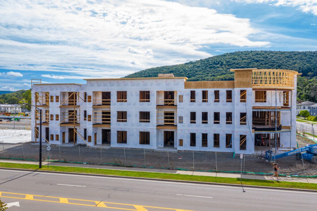 A three-story building under construction, with a white exterior wrap and wooden framing. Located near a road, with hills in the background. .