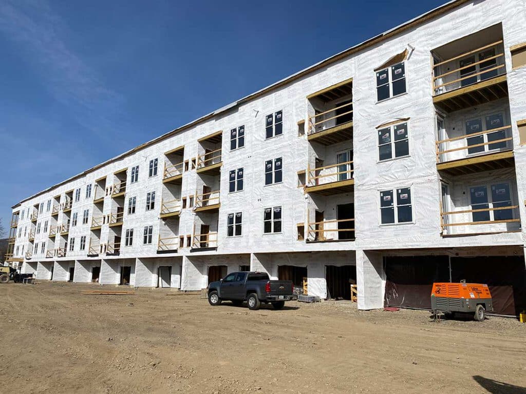 A large, unfinished multi-story building with white exterior wrap. Several balconies are visible. A truck and orange machine are parked in front, with dirt and clear sky in the background.