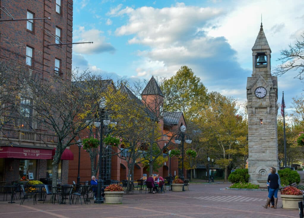 A public square with people sitting at tables, a brick building, and a stone clock tower under a cloudy sky.