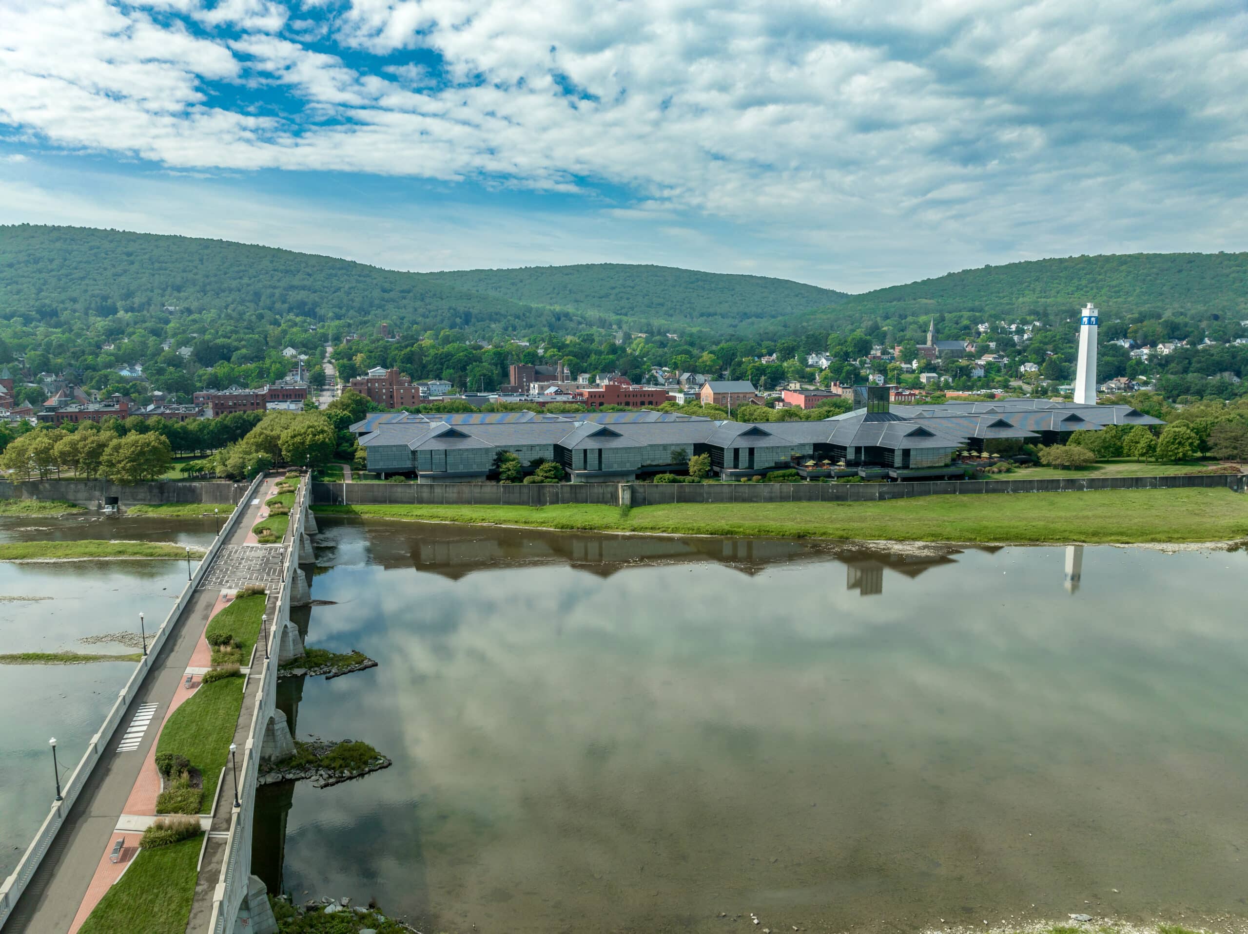 Aerial view of a large building complex near a body of water, surrounded by trees and hills under a partly cloudy sky. A bridge extends across the water in the foreground.