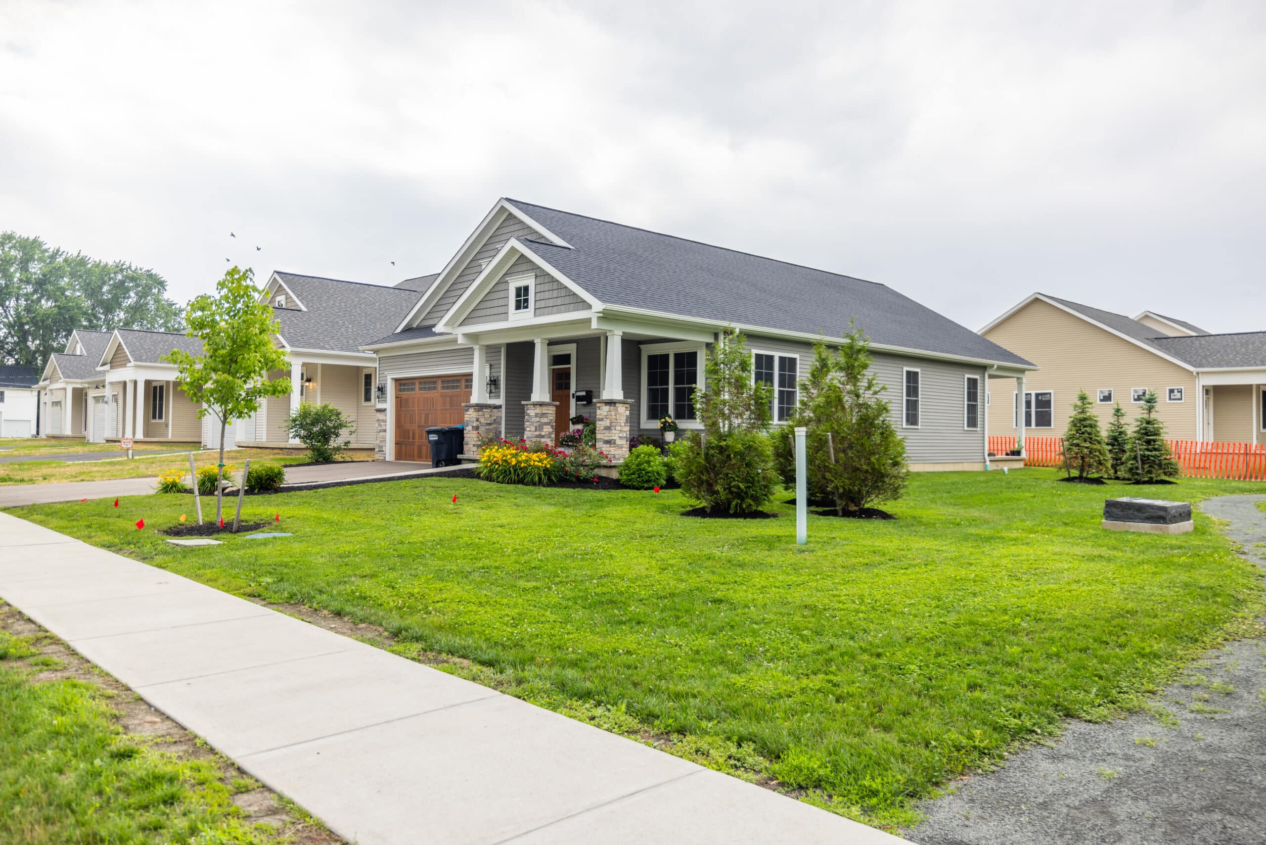 A single-story gray house with a front porch, surrounded by a well-maintained lawn and small trees. A sidewalk runs along the front. Cloudy sky overhead.