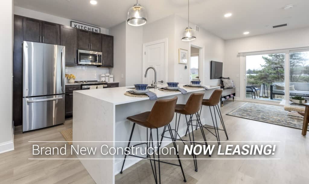 Modern kitchen with a white island, brown chairs, stainless steel appliances, and a view of a living area. Text overlay: "Brand New Construction! NOW LEASING!.