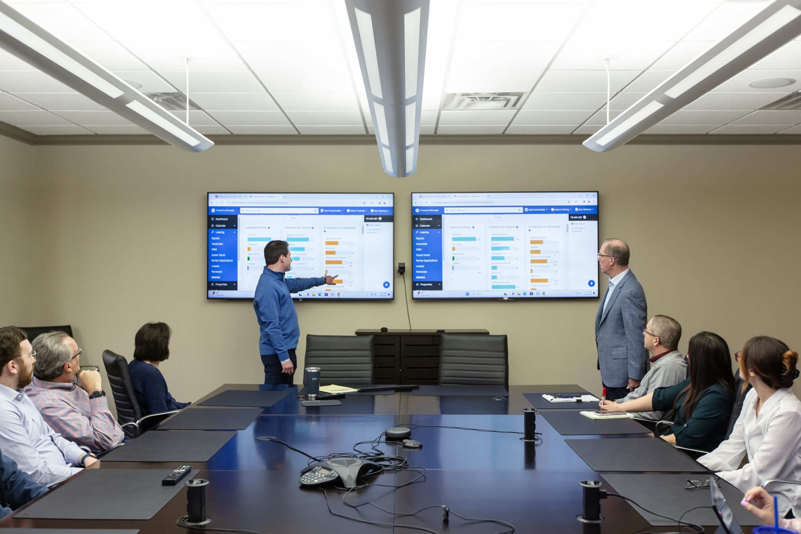 People in a conference room watch two men present data on two large screens.