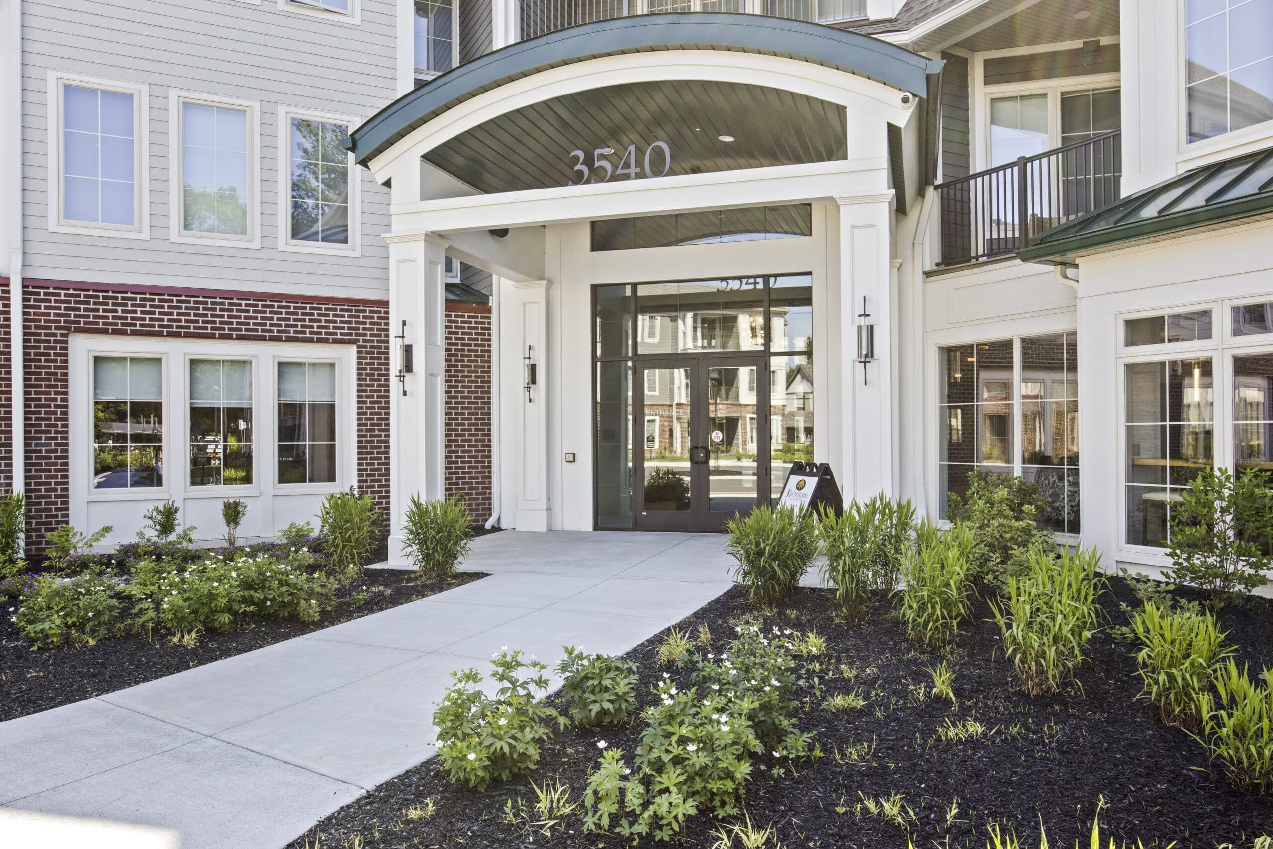 Entrance of a modern building with a glass door, overhang, and visible address number 3540. Surrounded by windows and landscaped garden.