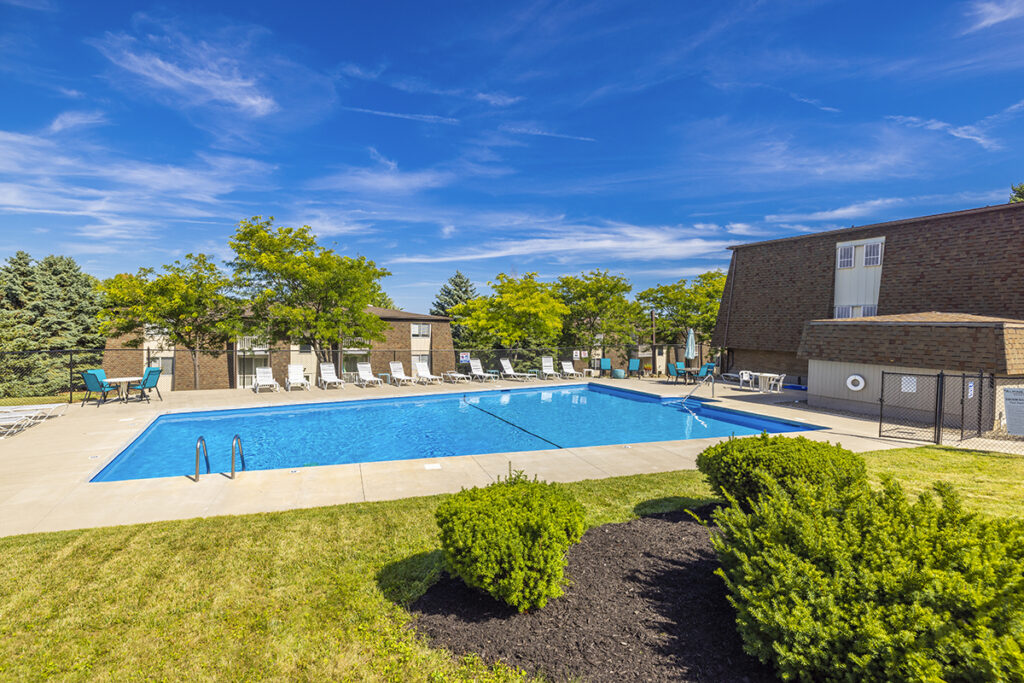 Inground pool at Alpine Village Apartments with a large sundeck, surrounded by mature landscaping. The fenced-in area offers plenty of lounge chairs and tables for residents to relax and enjoy the outdoor space. Ideal for unwinding on sunny days.