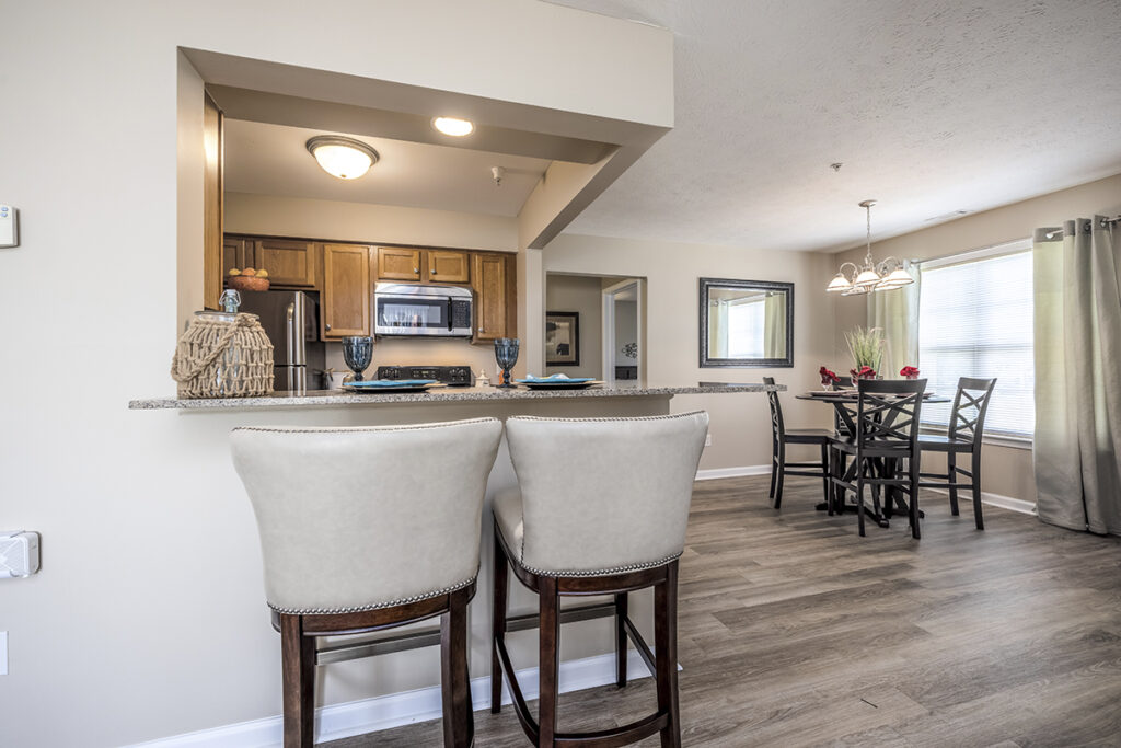 Bright dining room with a large window, luxury vinyl plank flooring, and a view of the breakfast bar in an open floor plan at The Hammocks at Orchard Park. Plenty of natural light fills the space.