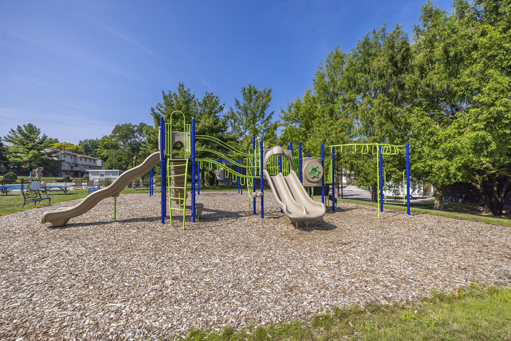 A playground with wood chips and seating, located adjacent to the pool area. The setup provides a safe and comfortable space for children to play while adults at a nearby bench.