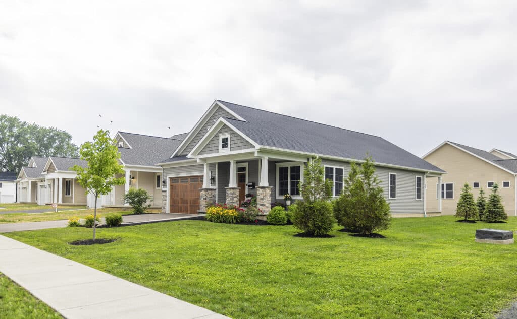 A single-story house with a gray roof and beige siding, surrounded by a well-maintained lawn and small bushes, under a cloudy sky.