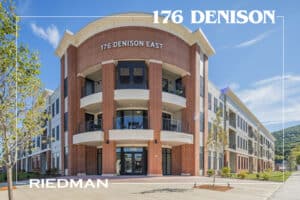 Brick and white modern three-story building with large windows and "176 Denison East" signage. Tree-lined walkway in front.