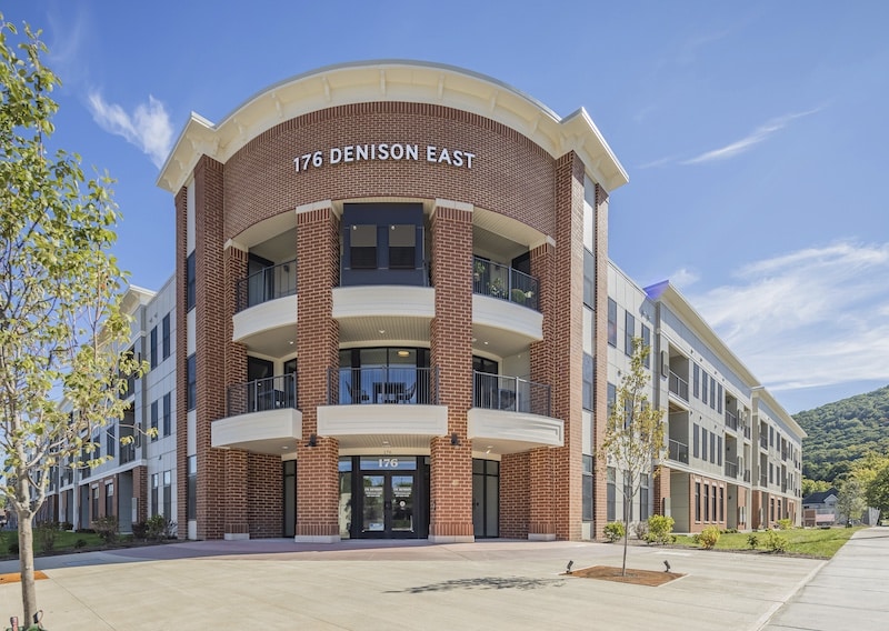 Three-story brick office building with white accents, labeled "176 Denison East," featuring multiple balconies and a central entrance. Trees and a clear sky surround the structure.