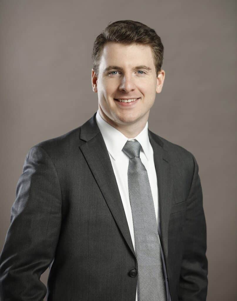 A man in a dark suit and tie stands against a neutral background, smiling at the camera.