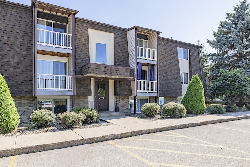 A two-story apartment building with a brown exterior and balconies. A paved walkway and parking area with accessible parking spaces are in the foreground. Trees and bushes are nearby.