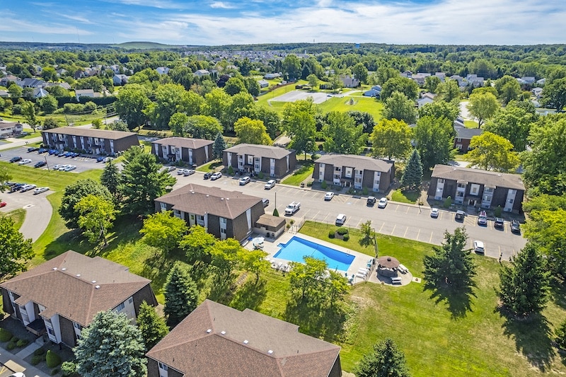Aerial view of a residential neighborhood with several apartment buildings, a swimming pool, parking areas, and surrounding greenery under a blue sky.