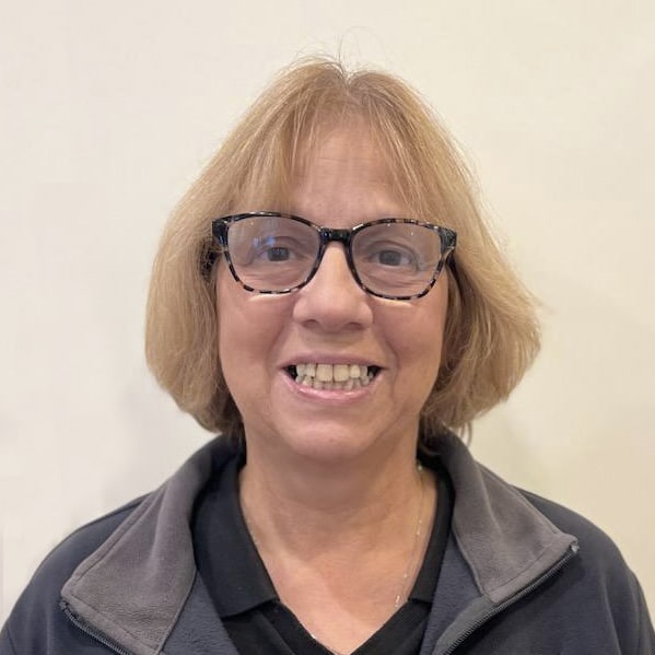 A person with short hair, wearing glasses and a zip-up jacket, smiles in front of a plain background.