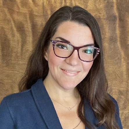 A person with long hair, wearing glasses and a blue blazer, smiles against a brown backdrop.