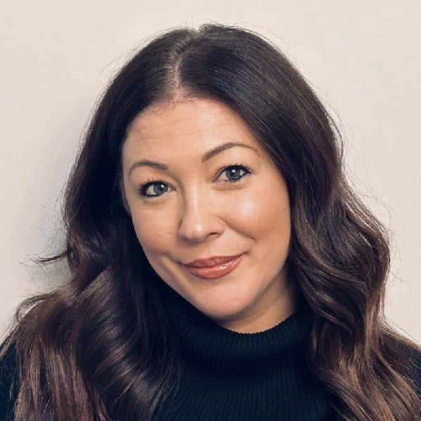 A person with long, wavy brown hair smiles slightly while wearing a dark sweater against a plain background.