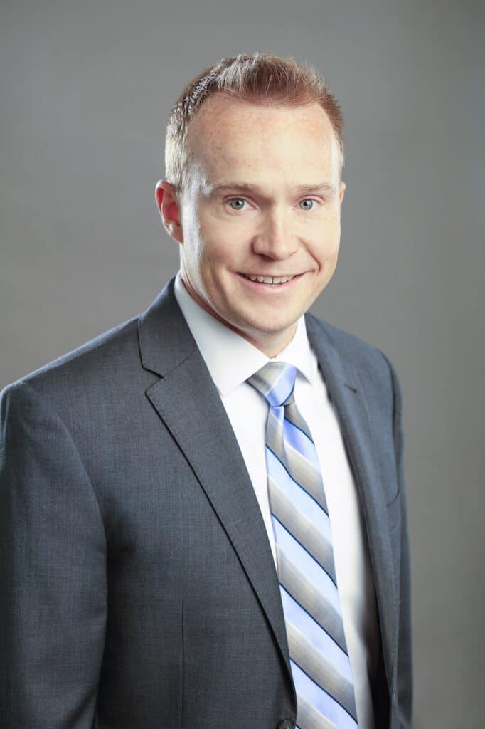 A man in a suit and tie smiles at the camera against a gray background.