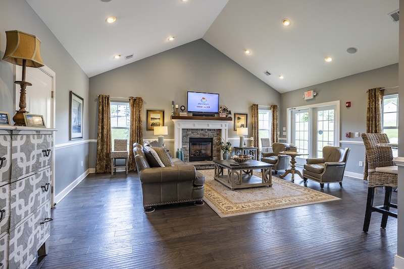 Spacious living room with gray walls, leather sofas, a fireplace, and a wall-mounted TV. Large windows and glass doors allow natural light. Neutral decor with wooden flooring and cushioned chairs.