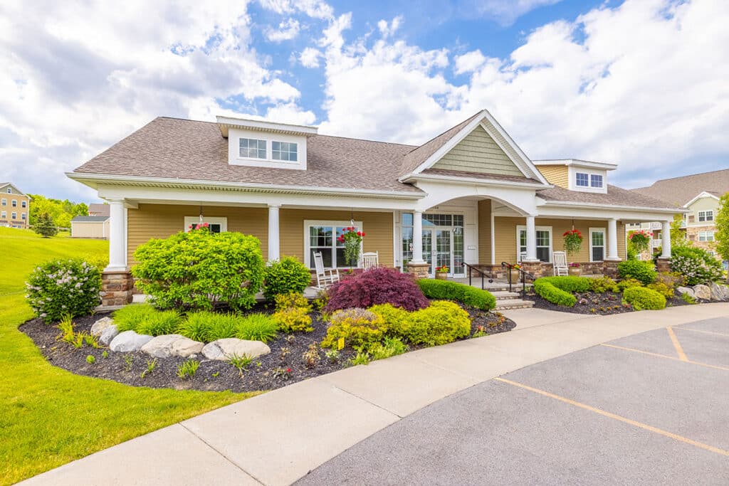 A single-story building with a gabled roof, large windows, and a landscaped front garden with shrubs and flowers, set against a partly cloudy sky.