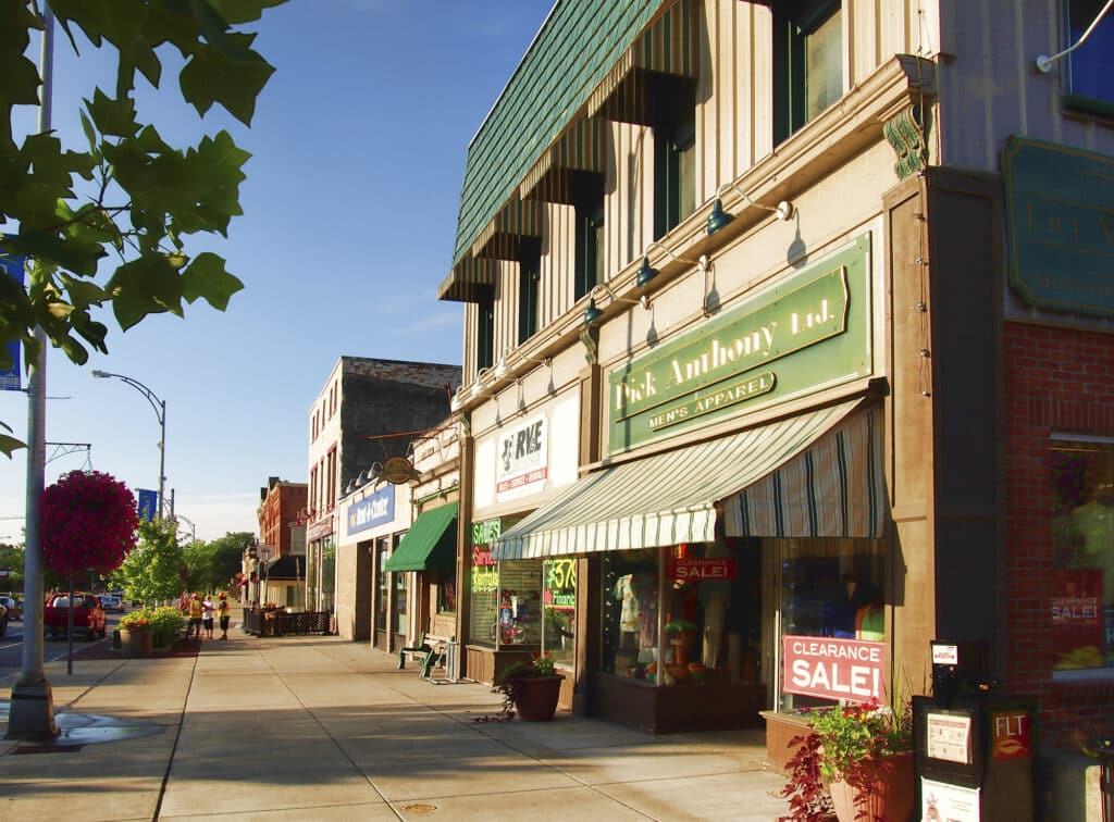 Street view of small-town shops, including men's apparel and clearance sale signs, lined along a sunny sidewalk.