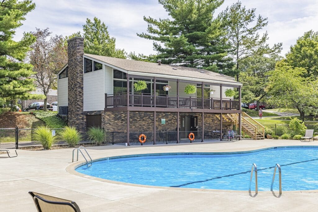 A two-story building with a pool in the foreground. The building has a brick lower level and a balcony with plants. Surrounded by trees, the scene is sunny and clear.