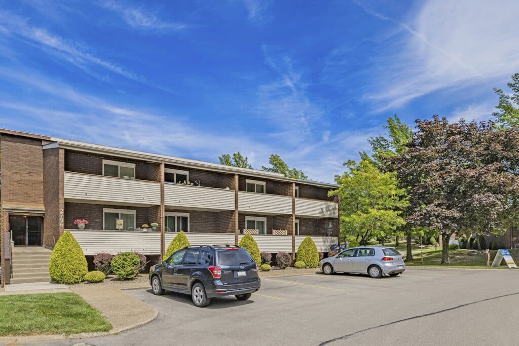 Two-story residential building with balconies, surrounded by trees and parked cars in front, under a clear blue sky.