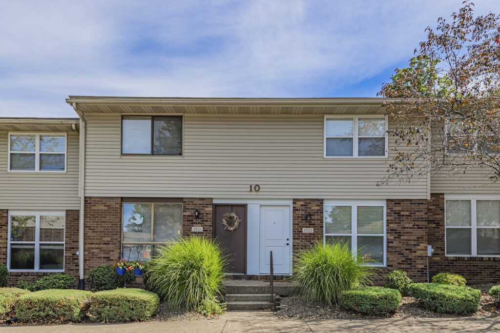 A two-story townhouse with beige siding and brick accents, featuring two entrance doors and windows, a wreath on one door, and bushes in front.