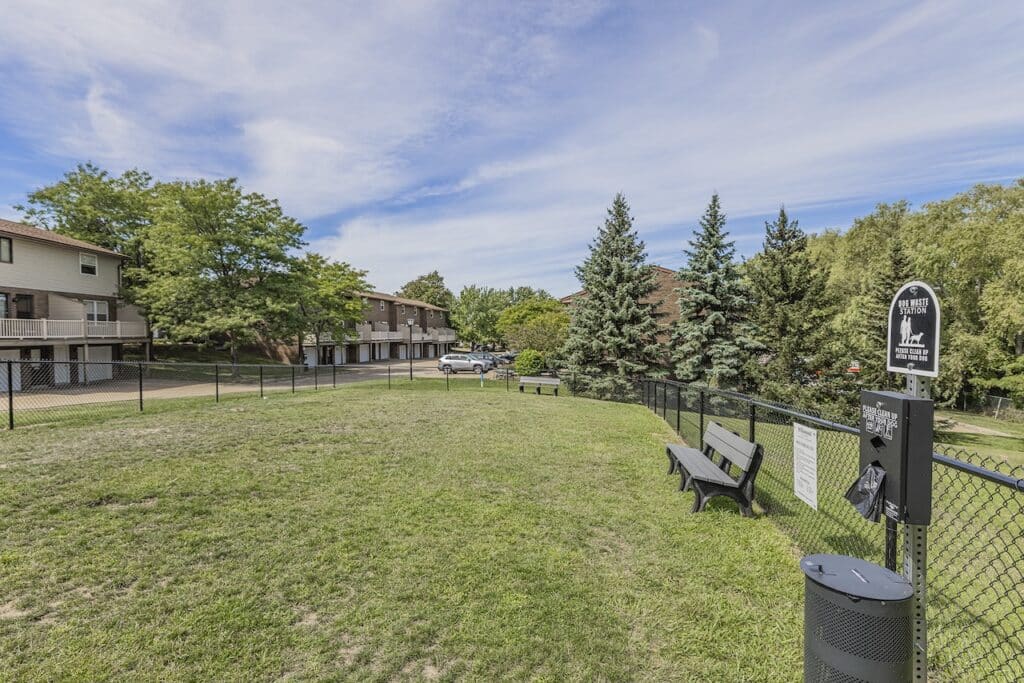 Dog park with benches, a waste station, and a surrounding fence. Nearby are residential buildings and trees under a blue sky.