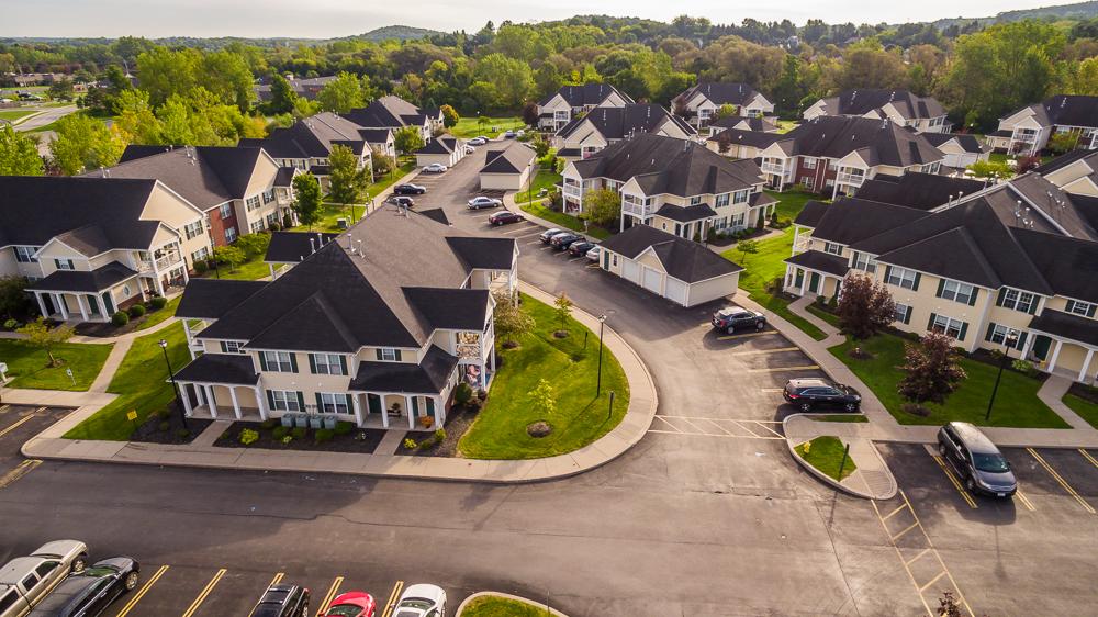 Aerial view of a suburban neighborhood with several houses, driveways, and parked cars. Green lawns and trees are visible throughout the area.