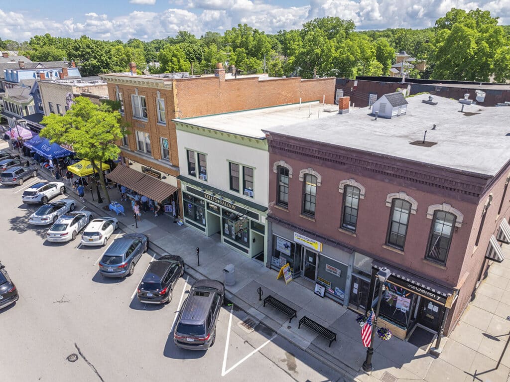 Aerial view of a small-town street with parked cars, brick buildings, trees, and clear blue sky overhead.