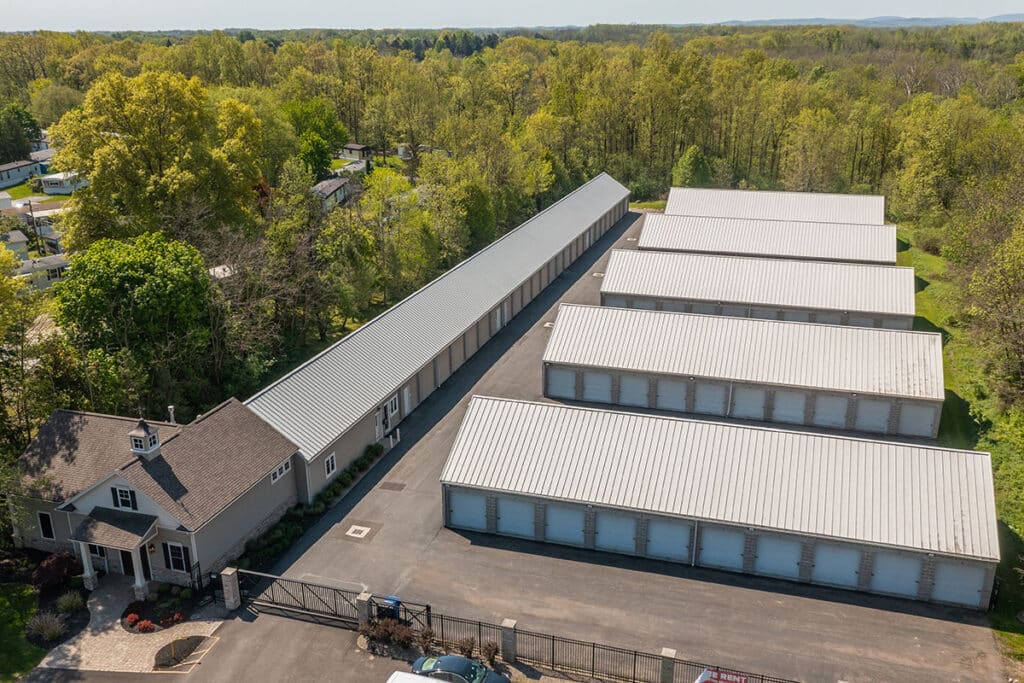 Aerial view of a storage facility with multiple large storage units and a small building at the front, surrounded by trees.