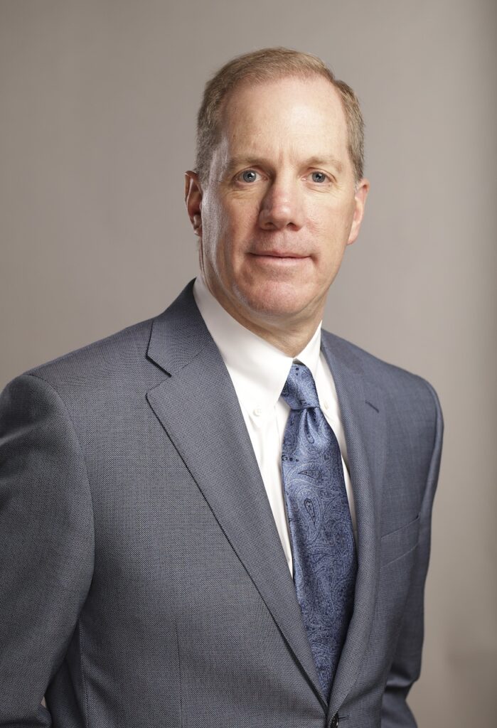 A man in a gray suit and blue tie stands against a neutral background.