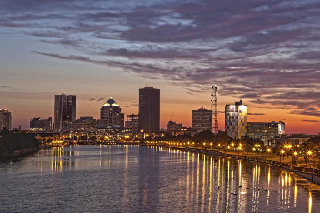 City skyline at sunset with buildings illuminated along a river, reflections visible on the water.