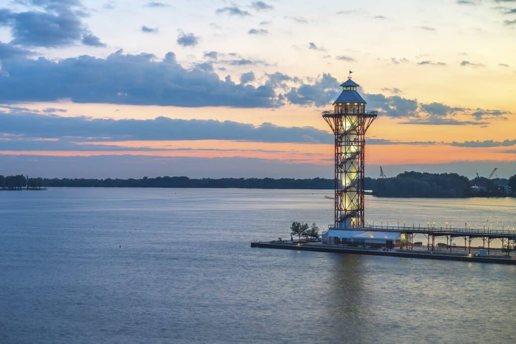 A lighthouse stands on a pier overlooking a calm body of water at sunset, with an orange sky and scattered clouds in the background.