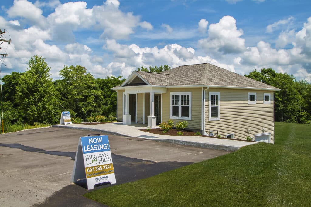 A small beige house with a "Now Leasing" sign and contact information in front, set against a backdrop of green trees and cloudy blue sky.