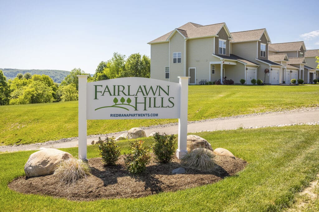 Sign for Fairlawn Hills apartments in front of a row of light green residential buildings, with manicured lawns and greenery in the background under a clear blue sky.