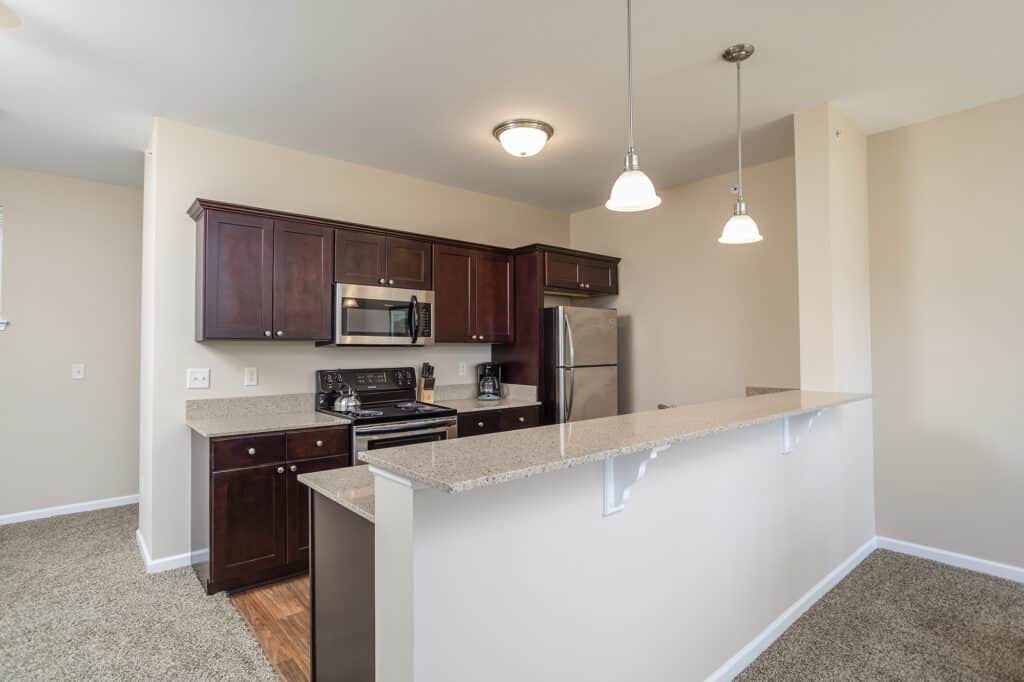 Modern kitchen with dark wood cabinets, stainless steel appliances, and a beige countertop bar. Carpeted flooring and pendant lights are present.