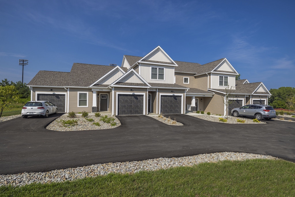 Building exterior view with sloped roofs, two garages, and parked cars on a paved driveway, under a clear blue sky.