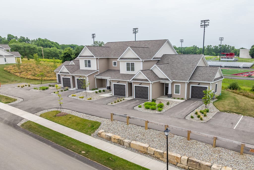 Attached townhomes with garages, landscaped yards, and a nearby sports field and bleachers in the background.