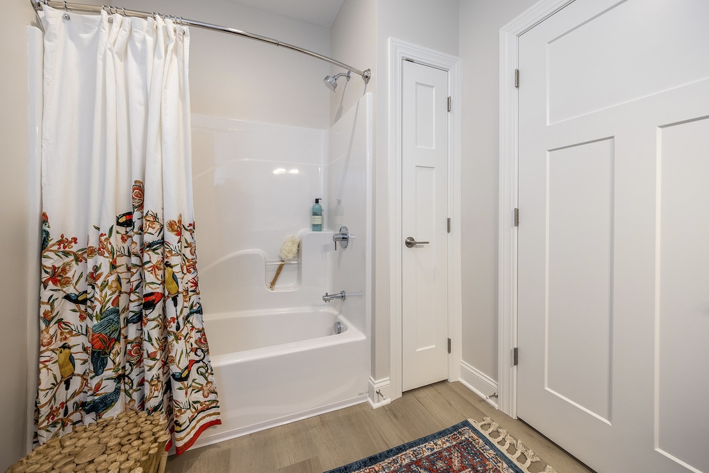 Bathroom with a bathtub, floral shower curtain, and a wooden stool. A bottle and scrub brush are on the bathtub ledge. White walls and door, with a patterned rug on the floor.