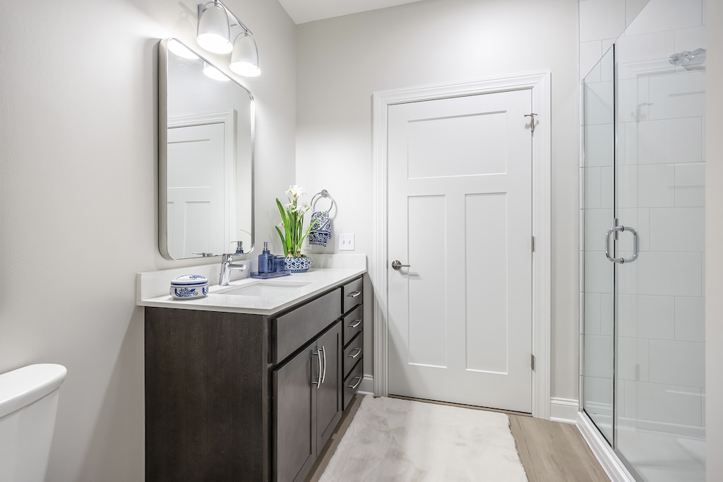 Modern bathroom with a dark wood vanity, white countertop, and vase with flowers. There's a large mirror, glass shower enclosure, and overhead lighting. The door is white.