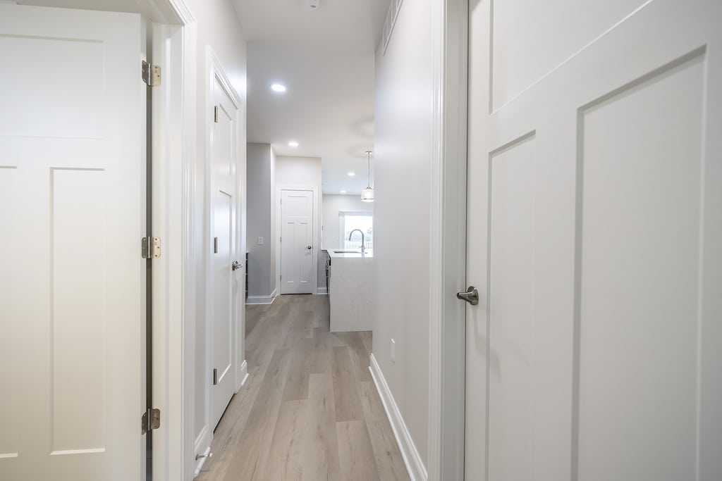 A bright, narrow hallway with light LVP flooring leads to a modern kitchen visible in the distance. White walls and doors line the corridor.