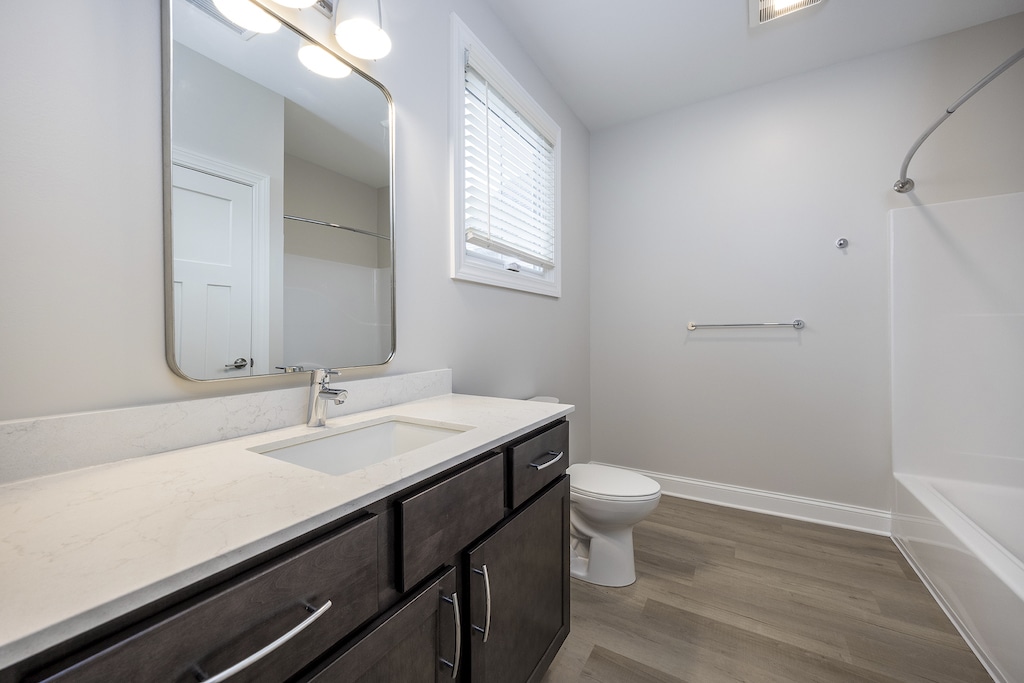 Bathroom with a large mirror above a sink on a dark maple vanity with quartz countertops, LVP flooring, toilet, bathtub with a shower curtain rod, and a frosted window.