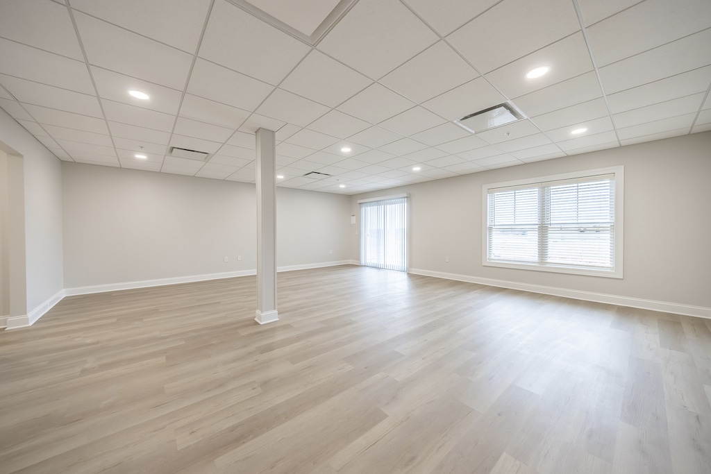 Finished walk-out basement with light LVP flooring, white walls, and a drop ceiling. Large window with blinds and a glass door letting in natural light.