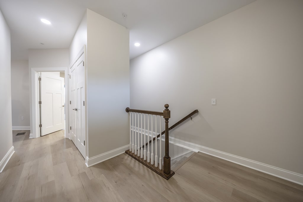 A hallway with light LVP flooring, white walls, and a staircase with a wooden railing leading to the downstairs main floor. There is a closed door and a small light illuminating the area.