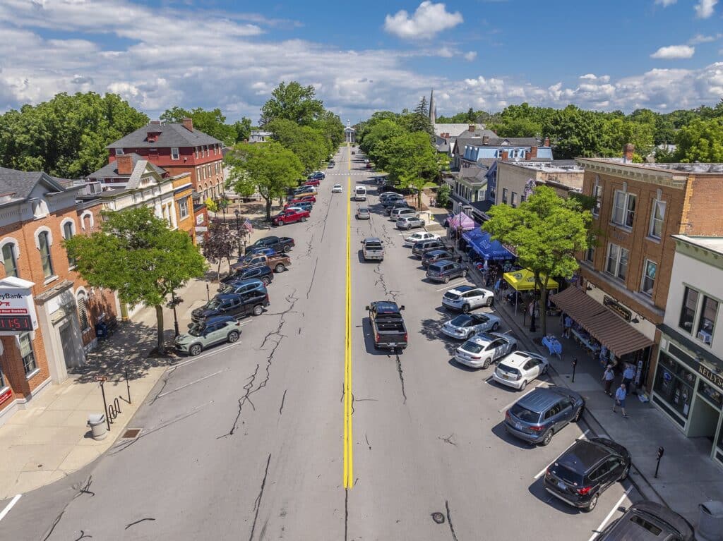 Aerial view of a small town street with cars parked along both sides, lined with brick buildings and trees under a partly cloudy sky.