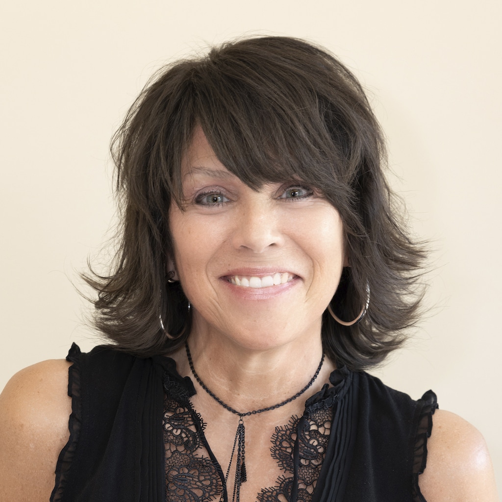 Smiling person with short, dark, layered hair and hoop earrings, wearing a black lace-trimmed top against a beige background.