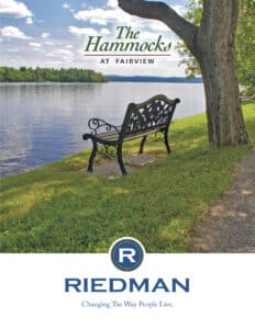 A metal bench on grass by a calm river with trees in the background. Text reads, "The Hammocks at Fairview" and "Riedman: Changing The Way People Live.